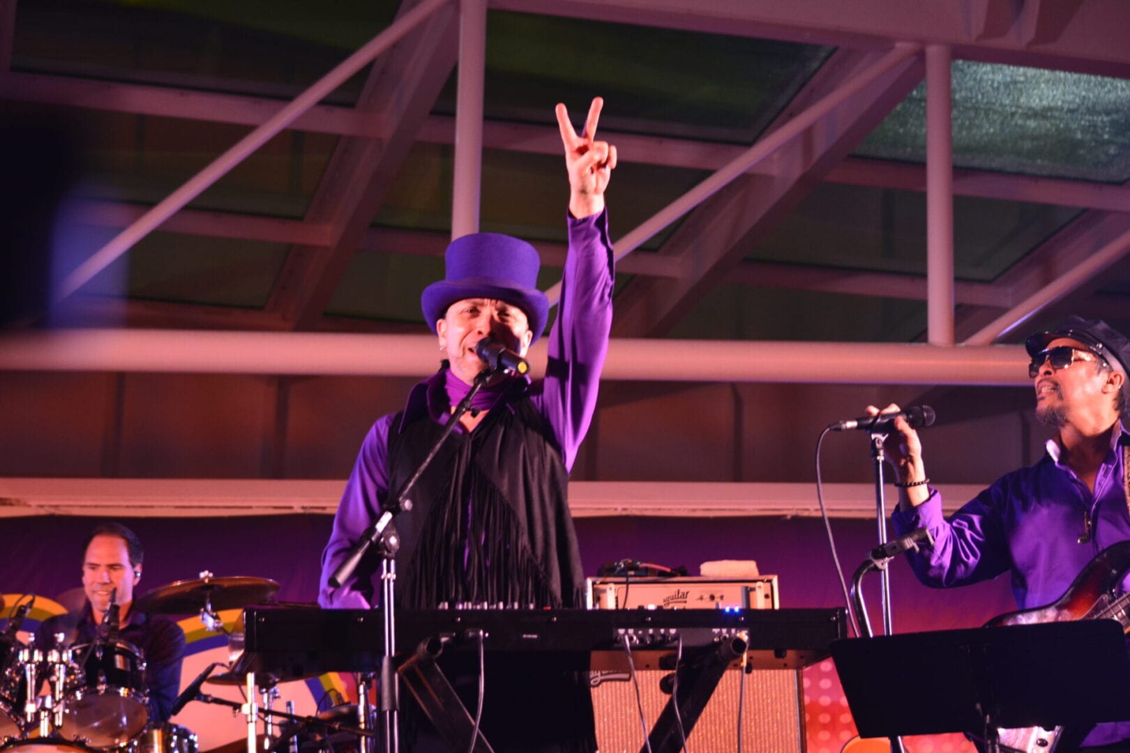A man in purple shirt and hat holding up peace sign.