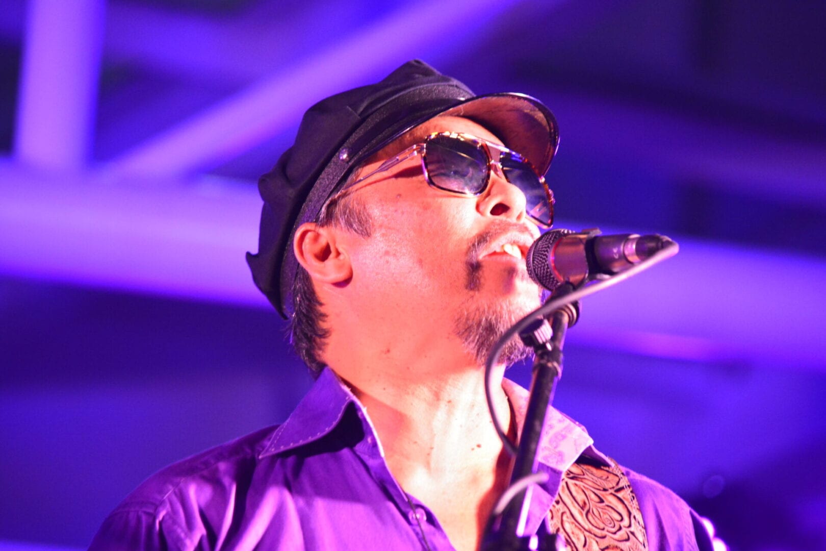 A man in purple shirt and hat singing into microphone.